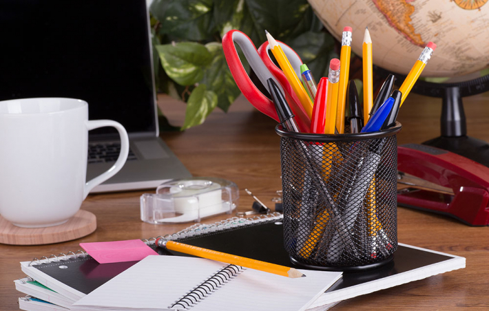 photo of a desk with a netted cup full of pens, pencils and a pair of scissors. there's a notebook underneath the cup with a few papers on top of that and a mug off to the left with a coaster underneath